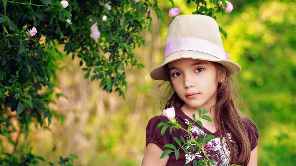 Free cute baby girl is having flower with leaf in hand wearing maroon top and light brown hat in green trees background hd cute wallpaper download