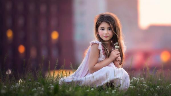 Free cute baby girl is sitting on green grass having flowers in hand wearing pink dress posing for a photo in blur background hd cute wallpaper download