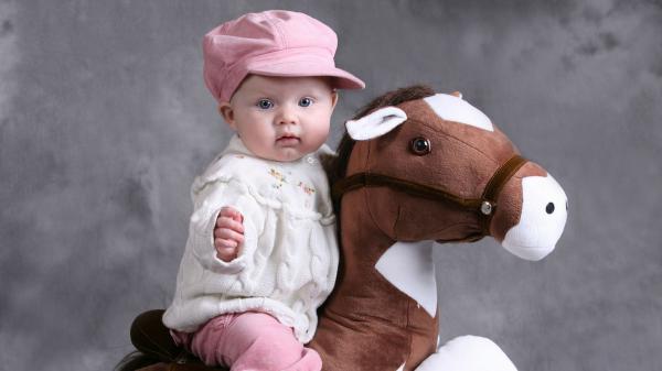 Free cute baby is sitting on toy horse wearing white top and pink pant and cap in ash background hd cute wallpaper download