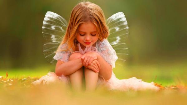 Free cute little girl is having wings on back looking down sitting on green grass wearing white dress during daytime hd cute wallpaper download