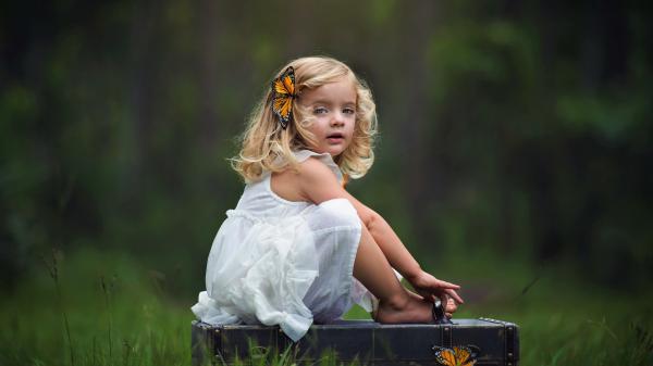 Free cute little girl is sitting on suitcase wearing white dress with butterfly clip in head in green blur background hd cute wallpaper download