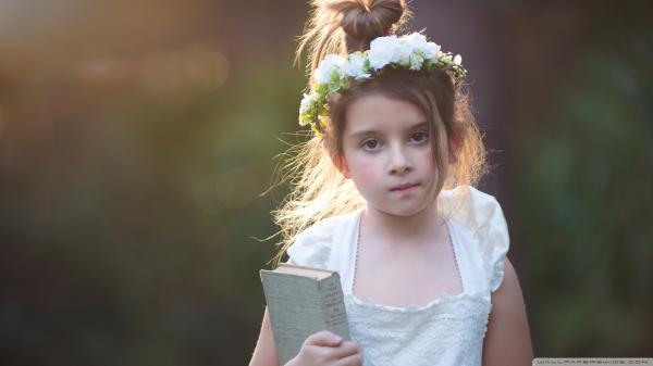 Free cute little girl is standing in blur background wearing white dress and flower crown on head holding book in hand hd cute wallpaper download