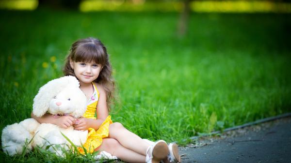 Free cute little girl is wearing yellow dress sitting on green grass in blur green background with teddy toy hd cute wallpaper download