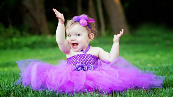 Free cute smiley girl baby is sitting on green grass wearing purple frock and flower band on head having hands in the air hd cute wallpaper download