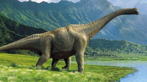 Free dinosaur is standing on grass field near water with mountain background hd dinosaur wallpaper download