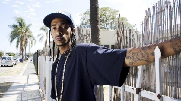 Free nipsey hussle is wearing blue tshirt having tattoos in hand standing in front of gate hd music wallpaper download