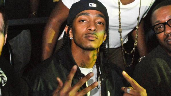 Free nipsey hussle is wearing white tshirt and black coat showing hand sign looking at camera hd music wallpaper download