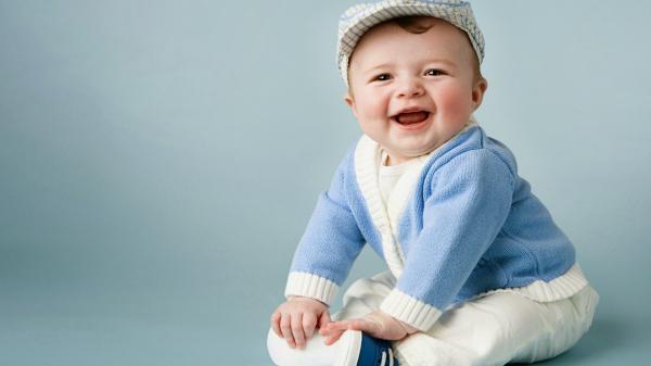 Free smiley cute baby boy is sitting on floor wearing white dress and cap with blue woolen sweater in blue background hd cute wallpaper download