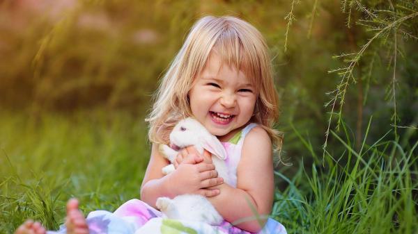 Free smiley cute baby girl is sitting on green grass holding rabbit wearing white dress in green blur background hd cute wallpaper download