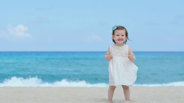 Free smiley cute girl baby is standing on beach sand wearing white dress in beach blur background hd cute wallpaper download