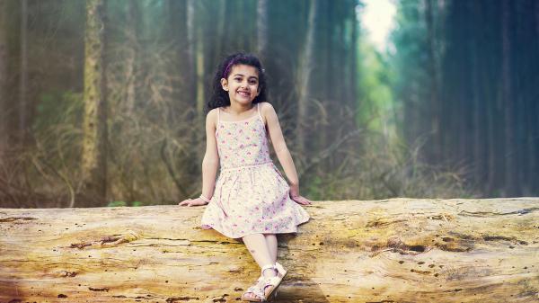 Free smiley cute girl is sitting on tree trunk wearing white flowers printed dress in forest background hd cute wallpaper download