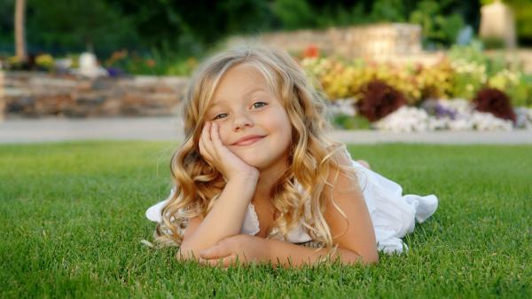 Free smiley cute little girl is lying down on green grass wearing white dress holding face in hand hd cute wallpaper download