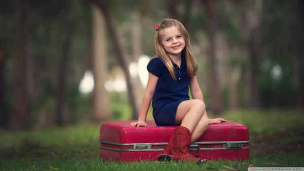 Free smiley cute little girl is wearing blue dress sitting on red suitcase in blur trees background hd cute wallpaper download