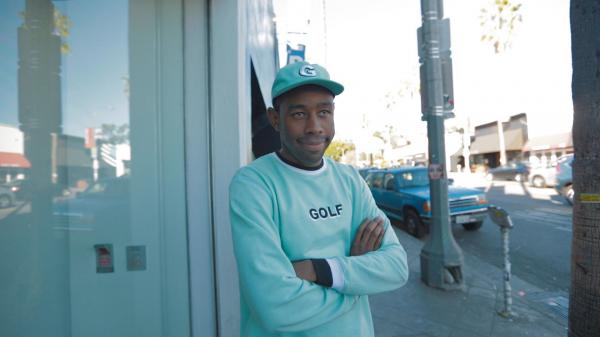 Free tyler the creator is smiling and looking straight standing near glass door wearing aquamarine tshirt and cap hd music wallpaper download